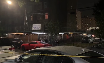 Noche en una calle de Brooklyn con coches estacionados y cinta policial amarilla en primer plano. Hay personas y edificios iluminados al fondo