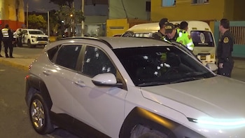 Automóvil SUV blanco con múltiples impactos de bala en los cristales y la carrocería. Agentes de policía inspeccionan el área en una calle urbana nocturna