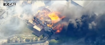 Vista aérea de una casa con fuego sobre el tejado y columnas densas de humo gris que cubren la propiedad y los alrededores