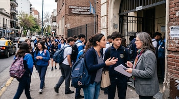Una mujer habla con una docente frente a la entrada de un colegio de ladrillo visto con alumnos uniformados de azul caminando por la vereda.