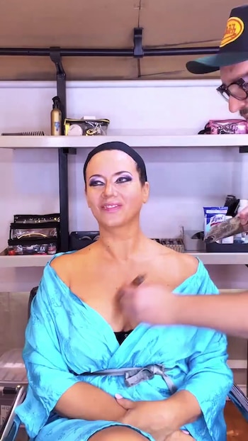Una mujer con una bata azul y una vincha negra recibe maquillaje en el cuello por parte de un maquillador en un vestidor