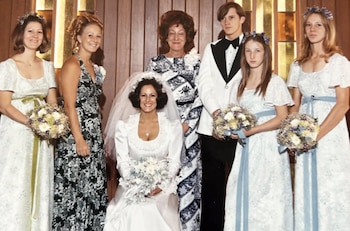 Una novia sonriente sentada en un vestido blanco con velo y ramo de flores, flanqueada por seis asistentes, incluyendo un hombre de esmoquin y cinco mujeres con vestidos florales y ramos