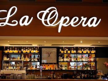 Cartel luminoso con la palabra 'La Opera' sobre una barra de bar con estantes llenos de botellas, copas, vasos y platos apilados