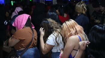 Un grupo de mujeres en un lugar oscuro y concurrido. Una mujer cubre su cabeza con una tela rosa y otra su rostro con la mano. Se observan personas y policías