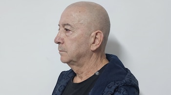 Hombre calvo de perfil izquierdo, viste camiseta negra y chaqueta acolchada azul marino sobre fondo blanco.