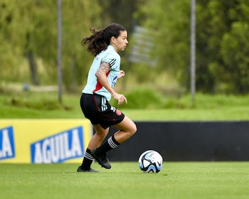 Una futbolista con camiseta celeste y pantalones cortos negros conduce un balón en una cancha de césped