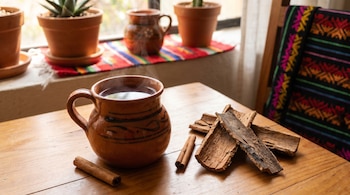 Una jarra de barro con té caliente, corteza seca, ramas de canela, dos macetas con plantas, un mantel y una silla decorada sobre una mesa de madera.