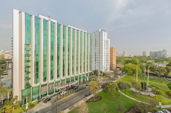 Vista exterior elevada del Delfines Hotel, un edificio alto con fachada de vidrio verde y blanco, junto a una calle y un parque con árboles