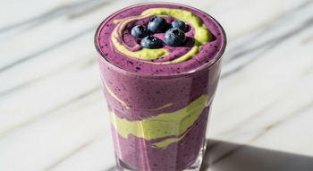 Primer plano de un vaso con batido de color morado y verde, decorado con arándanos, sobre una superficie de mármol blanco.