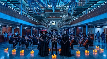 Tres personas con trajes negros y maquillaje de calavera frente a una orquesta, velas en el suelo y estanterías iluminadas con luz azul.