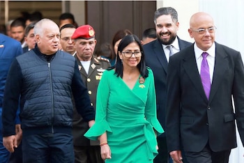 Jorge Rodríguez con chaleco oscuro y Delcy Rodríguez con vestido verde y gafas, caminan sonriendo junto a otros hombres en trajes y un militar en uniforme
