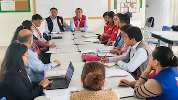 Un grupo de personas sentadas en una reunión alrededor de una mesa con documentos. Hay un portátil y teléfonos celulares. Algunas personas visten chalecos rojos
