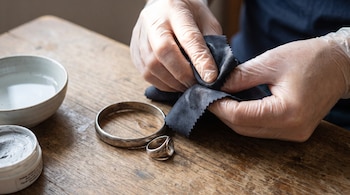 Manos enguantadas sostienen un paño oscuro. En una mesa de madera hay una pulsera de plata, un anillo de plata, un cuenco con líquido y un envase de pasta limpiadora.