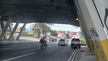 Automóviles, una camioneta y una motocicleta avanzan por una carretera bajo una estructura de puente de concreto con grafiti