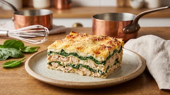 Porción de lasaña en plato, capas de pollo desmenuzado, espinaca, bechamel y queso gratinado. Fondo con dos cazos de cobre, batidor y hojas de espinaca.