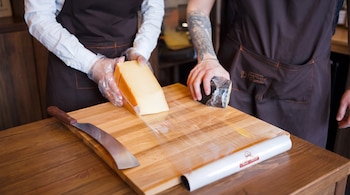 Dos personas con delantales y guantes de plástico manipulan dos bloques de queso, uno amarillo y otro oscuro, sobre una tabla de madera con film transparente y un cuchillo.