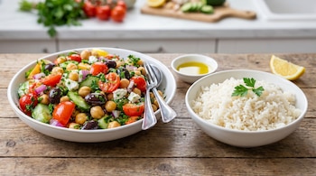 Un tazón de ensalada griega con garbanzos, tomates, pepino, aceitunas y queso. Un tazón de arroz. Aceite de oliva y una rodaja de limón.