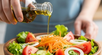 Primer plano de una mano vertiendo un chorro de aceite de oliva desde una botella de vidrio sobre una ensalada fresca con lechuga, tomate, zanahoria y cebolla.