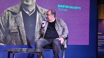 Un hombre de mediana edad con gafas y camisa a cuadros, sentado en una silla negra, habla por un micrófono con el logo de Ticmas frente a una pantalla morada