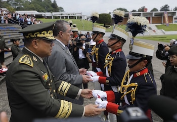 Oficial del ejército y civil intercambian objetos con dos candidatos en uniforme ceremonial con sombreros de plumas. Otros militares y un edificio de escuela al fondo