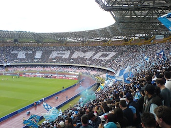 Estadio de fútbol lleno de público con banderas azules y blancas. Personas en las gradas forman la frase "Ti amo". Césped, pista de atletismo y estructura del techo