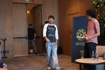 Louis Tomlinson de pie, sonriendo, vestido con camiseta blanca y oscura, y jeans rasgados, en una sala con un banner de BMG y una planta grande