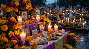 Altar con flores naranjas y moradas, velas encendidas, pan, calaveras y retratos enmarcados frente a tumbas iluminadas por velas