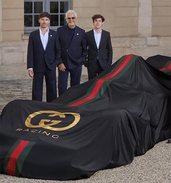 Tres hombres con trajes oscuros posan frente a una fachada de piedra, junto a un coche cubierto por una tela negra con el logo dorado de Gucci Racing y franjas rojas y verdes