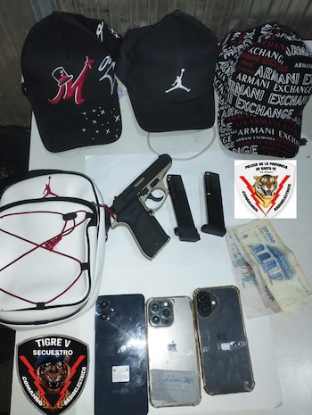 Vista aérea de tres gorras, una cartera blanca tipo bandolera, una pistola, dos cargadores, billetes, tres teléfonos celulares y dos insignias policiales sobre una superficie clara
