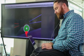 Un hombre con barba usa una computadora portátil frente a una pantalla grande con gráficos digitales tridimensionales