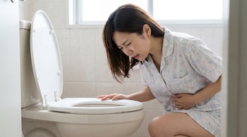 Mujer japonesa en pijama claro, agachada frente a un inodoro blanco con la tapa levantada, se sujeta el abdomen con expresión de dolor y náuseas.