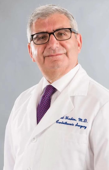 Un hombre de gafas y cabello canoso viste una bata blanca de laboratorio con bordado azul, camisa blanca y corbata estampada