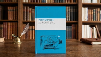 Libro de tapa dura azul titulado 'Un inmenso azul' de Patrik Svensson, con ilustración de un barco en una botella, sobre un escritorio de madera; al fondo, una biblioteca con libros