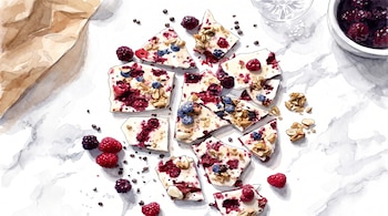 Trozos de chocolate blanco o yogur con frambuesas, arándanos, moras y granola esparcidos sobre una mesa de mármol; un bol con bayas y un vaso a la derecha.