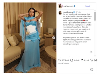Maria Becerra de pie con vestido azul y blanco y broche de sol. Fondo con sofá beige y cortinas. Captura de pantalla de publicación de Instagram