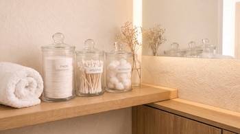 Tres frascos de vidrio con etiquetas de texto, una toalla blanca enrollada y un florero pequeño sobre un estante de madera frente a un espejo.