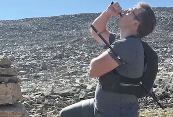 Hombre con gafas bebe de una manguera de hidratación. Lleva mochila y un bastón de trekking. Alrededor hay rocas, un cielo azul y un apilamiento de piedras