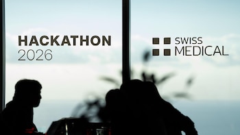 Texto 'HACKATHON 2026' y 'SWISS MEDICAL' con logo en un cristal, con siluetas borrosas de personas en primer plano y cielo de fondo