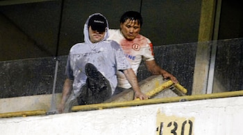 Dos hombres en un palco de estadio. Uno viste sudadera gris y gorra negra. Otro lleva camiseta blanca con logo U y manchas rojas. Hay una baranda y el número 130