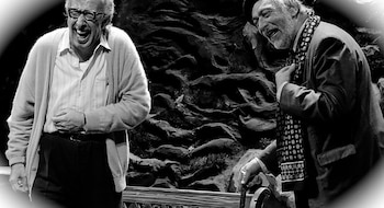 Fotografía en blanco y negro de Luis Brandoni y Eduardo Blanco riendo. Brandoni, a la izquierda, con camisa y cárdigan. Blanco, a la derecha, con traje y bufanda