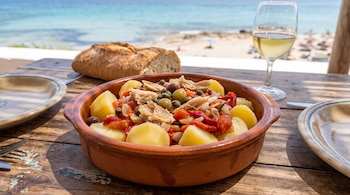Cuenco de ensalada de patatas, pimientos, pescado y aceitunas, con pan y copa de vino sobre mesa de madera rústica frente a playa y mar azul.