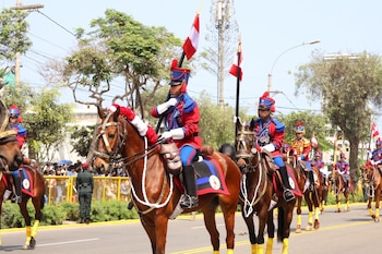 Grupo de jinetes militares en uniformes rojos y azules sobre caballos marrones, portando banderas de Perú. Calle arbolada con público detrás de vallas