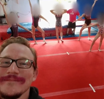 Un hombre con gafas se toma una fotografía en un gimnasio frente a varias jóvenes gimnastas con los rostros difuminados