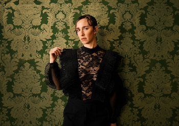 Mujer, blusa negra de encaje, volantes, pantalón negro, cabello corto peinado, fondo verde con estampado ornamental