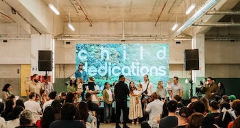 Un grupo diverso de adultos y niños se encuentra en un escenario frente a una gran pantalla con el texto 'child dedications', mientras una audiencia los observa