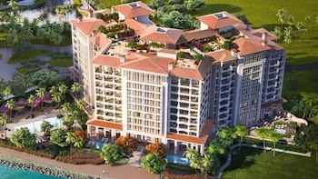 Vista aérea de un complejo hotelero con edificios de color claro, techos de terracota, balcones, piscinas y palmeras junto a un cuerpo de agua