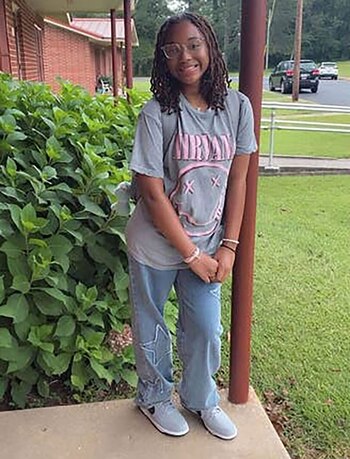 Jada West, una joven con dreadlocks y gafas, sonríe ligeramente con las manos juntas frente a un edificio de ladrillo, vegetación y una carretera