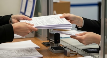 Primer plano de un par de manos entregando una pila de documentos a otro par de manos en una ventanilla. Un escritorio con formularios y sellos es visible.