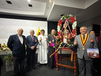 Grupo de cinco hombres, incluyendo religiosos, sonríe junto a la estatua de San Pedrito adornada con flores rojas y blancas, en un evento con pantalla y bandera