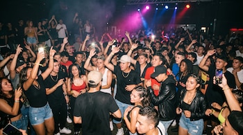 Decenas de jóvenes bailan en una discoteca iluminada con luces de colores mientras algunos sostienen teléfonos móviles con la mano levantada.